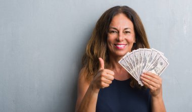 Orta yaş İspanyol kadın dolar ile mutlu büyük holding gri grunge duvardan ayakta gülümseme ok işareti, başparmak ile parmak, mükemmel işareti yapıyor