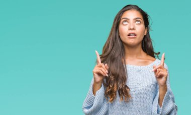 İzole arka plan üzerinde kış kazak giyen genç güzel Arap kadın hayrete ve yukarıya bakıyor ve parmakları ile işaret ve silah kaldırdı şaşırttı.