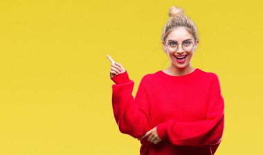 Kameraya bakarak yan el ve parmak ile işaret yüzünde kocaman bir gülümseme ile izole arka plan üzerinde kırmızı kazak ve gözlük takan genç güzel sarışın kadın.