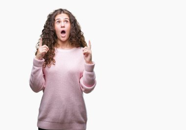 İzole arka plan üzerinde pembe kış kazak giyen genç kız hayran ve yukarıya bakıp parmakları ile işaret şaşırttı ve silah kaldırdı güzel esmer kıvırcık saç.