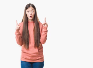 Genç Çinli kadın giyiyor spor sweathshirt hayret ve yukarıya bakıp parmakları ile işaret şaşırttı ve silah kaldırdı izole arka plan üzerinde.