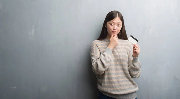 年轻的中国女人过了灰色的墙拿着信用卡严肃的面对思考的问题 很迷茫的想法 — 图库照片