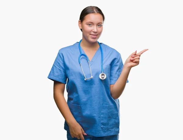 若い白人の医者の女性の顔に大きな笑みを浮かべて隔離された背景に医療制服を着てカメラを見て側に手と指で指しています — ストック写真
