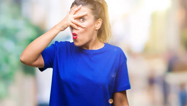 Young beautiful woman wearing casual blue t-shirt over isolated background peeking in shock covering face and eyes with hand, looking through fingers with embarrassed expression.