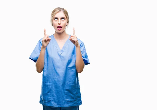 年轻美丽的金发医生外科医生护士妇女在孤立的背景惊讶和惊讶地抬起头来 用手指指指点点 举起双臂 — 图库照片