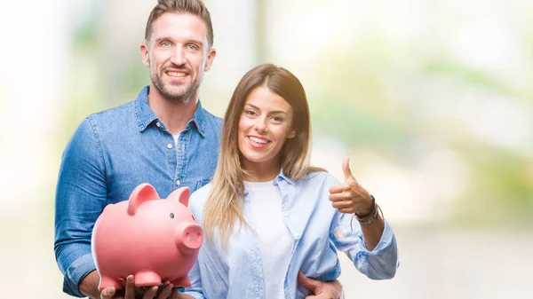 Young couple in love holding piggy bank over isolated background happy with big smile doing ok sign, thumb up with fingers, excellent sign