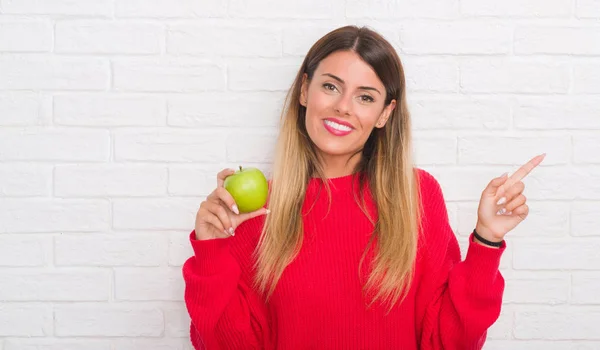 年轻的成年妇女在白色砖墙吃新鲜的绿色苹果非常高兴用手和手指指着一边 — 图库照片