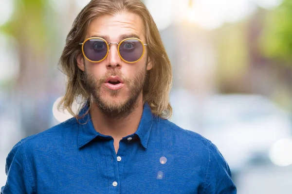 Korkuyor ve şok sürpriz ifade, korku ve heyecan yüz ile izole arka plan üzerinde güneş gözlüğü takmış uzun saçlı yakışıklı delikanlı.