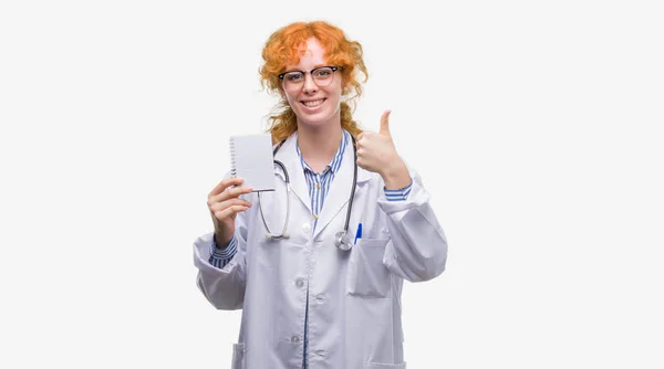 赤毛の若い医師女性と幸せが大きな空白のノートブックを保持して笑顔 サイン 優秀な兆候を親指をやって — ストック写真