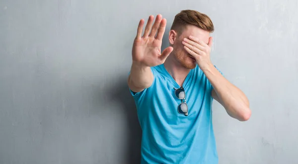 Young redhead man over grey grunge wall wearing casual outfit covering eyes with hands and doing stop gesture with sad and fear expression. Embarrassed and negative concept.