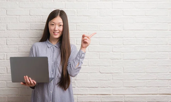 年轻的中国女人在砖墙上使用笔记本电脑非常高兴用手和手指指着一边 — 图库照片