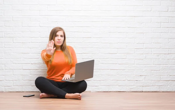 年轻的成年妇女坐在地板上 在白色砖墙上使用电脑笔记本电脑与开放的手做停止标志与严肃和自信的表情 防御手势 — 图库照片