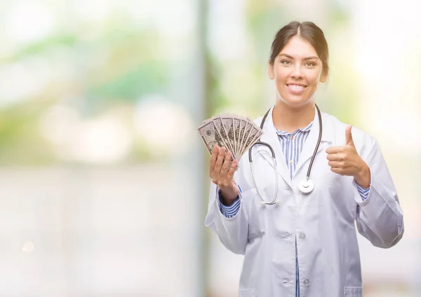 Young hispanic doctor woman holding dollars happy with big smile doing ok sign, thumb up with fingers, excellent sign