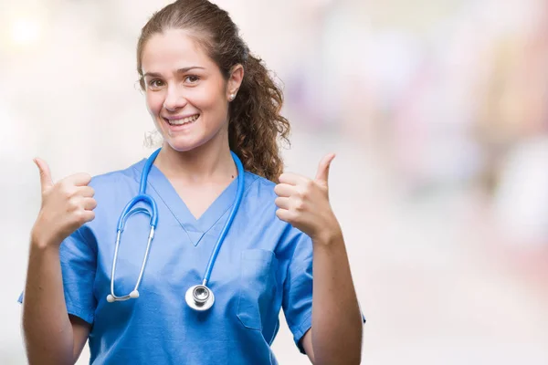 年轻的黑发医生女孩穿着护士或外科医生制服在孤立的背景成功标志做积极的手势与手 竖起大拇指微笑和快乐 带着欢快的表情 获胜者的手势看着镜头 — 图库照片