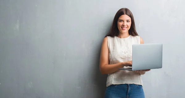 年轻的黑发女人在粗糙的灰色墙壁上使用电脑笔记本电脑与一个快乐的脸站着 面带微笑 自信的微笑显示牙齿 — 图库照片