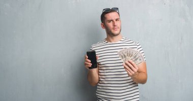 Beyaz delikanlı yüz çapraz silah ile dolar ve smarphone şüpheci ve sinir, onaylamayan ifade tutan gri grunge duvarın üzerinden. Negatif kişi.