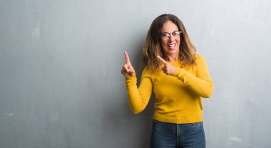 Orta yaş İspanyol kadın gülümseyerek ve iki elleri ve parmakları ile tarafına işaret kamera bakarak gözlüklü gri duvarın üzerinden.