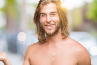 Genç yakışıklı gömleksiz adam uzun saçlı seksi vücut neşeli sunulması ve kameraya bakarak avucunun ile işaret gülümseyen izole arka plan üzerinde gösterilen.