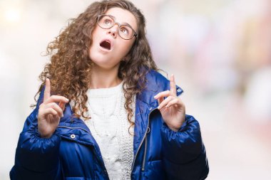 Kış ceket, gözlük ve kazak izole arka plan üzerinde giyen güzel genç esmer kıvırcık saçlı kız hayran ve yukarıya bakıp parmakları ile işaret şaşırttı ve silah kaldırdı.