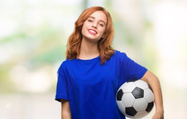 İzole arka plan ayakta ve diş gösterilen kendine güvenen bir gülümsemeyle gülümseyerek mutlu bir yüz ile futbol futbol topu tutan genç güzel kadın