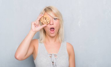 Gri grunge duvar yeme üzerinde beyaz yetişkin kadın çikolata bisküvi bir sürpriz yüz, korkuyor ve heyecanlı korku ifadesi ile şok korkmuş