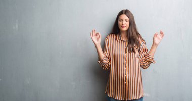 Genç esmer kadın grunge gri duvarın üzerinden rahatla ve gülümseyen gözlerle parmak ile meditasyon jest yapıyor kapalı. Yoga kavramı.