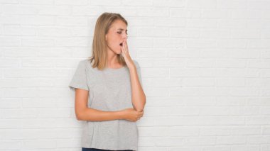 Güzel genç kadın beyaz tuğla duvarın üzerinden el ile ağız kapsayan yorgun esneme sıkılmış. Restless ve uyku hali.