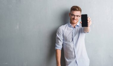 Gri grunge duvar gösteren boş ekran smartphone ayakta ve diş gösterilen kendine güvenen bir gülümsemeyle gülümseyerek mutlu bir yüz ile genç Kızıl saçlı adam