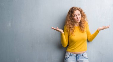 Gri grunge duvar clueless ve şaşkın bir ifadeyle kollar ve eller kaldırdı genç Kızıl saçlı kadın. Şüphe kavramı.