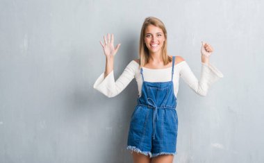 Gösterilen ve parmakları ile işaret grunge gri duvar üzerinde duran güzel genç kadın kendinden emin ve mutlu gülümseyerek süre 6 numara.