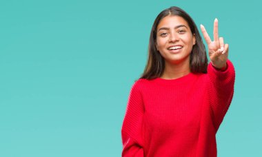 Zafer işareti yapıyor parmak gösterilen kamera seyir gülümseyen izole arka plan üzerinde kış kazak giyen genç güzel Arap kadın. İki numaralı.