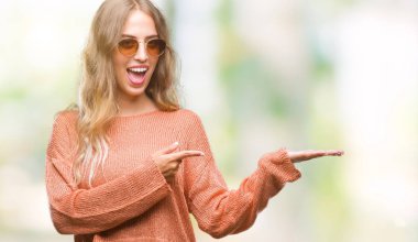 Güzel genç sarışın kadın hayrete ve el ile sunulması ve parmak ile işaret ederken kameraya gülümseyen izole arka plan üzerinde güneş gözlüğü takıyor.