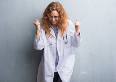 Gülümseyen ve başarı için çığlık genç redhead doktor kadını çok mutlu ve heyecanlı kaldırdı, silah ile kazanan jest yapıyor bir palto giyiyor gri grunge duvarın üzerinden. Kutlama kavramı.