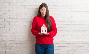 Genç Çinli gerçek durum Aracısı kadın ayakta ve diş gösterilen kendine güvenen bir gülümsemeyle gülümseyerek mutlu bir yüz ile ev tutan tuğla duvarın üzerinden