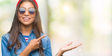 Genç güzel Arap kadın hayrete ve el ile sunulması ve parmak ile işaret ederken kameraya gülümseyen izole arka plan üzerinde güneş gözlüğü takıyor.