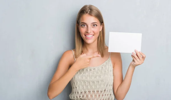 非常に幸せな手と指で指している空白のカードを保持しているグランジ灰色の壁に美しい若い女性 — ストック写真