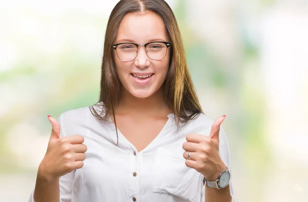 Young caucasian beautiful business woman wearing glasses over isolated background success sign doing positive gesture with hand, thumbs up smiling and happy. Looking at the camera with cheerful expression, winner gesture.