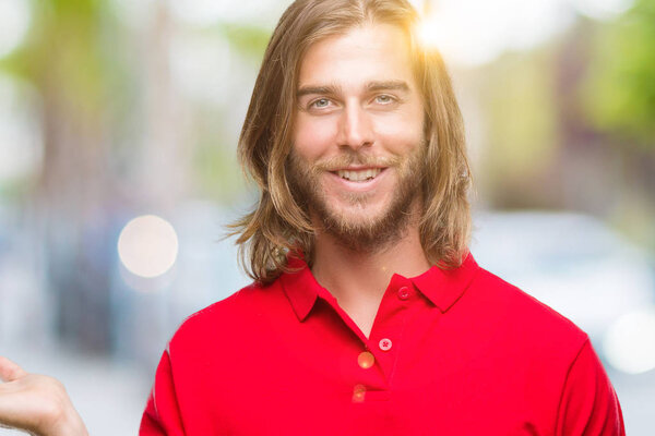 Young handsome man with long hair over isolated background smiling cheerful presenting and pointing with palm of hand looking at the camera.