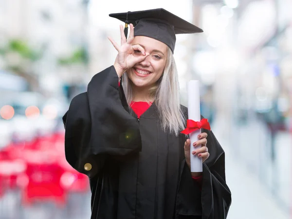 指の間から見て目に手で サインをして笑っている幸せそうな顔で孤立の背景の上の学位を保持する大学院の制服を着て若いブロンドの女性 — ストック写真