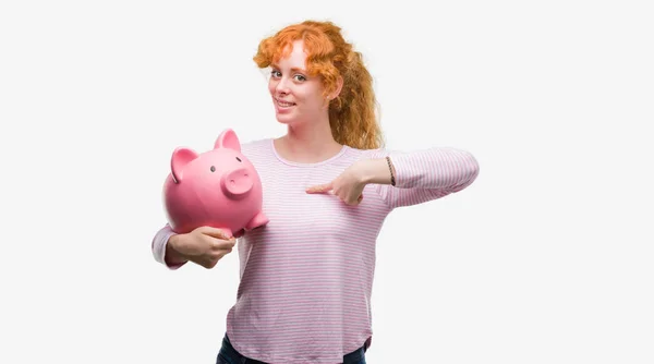 Young Redhead Woman Holding Piggy Bank Surprise Face Pointing Finger — Stock Photo, Image