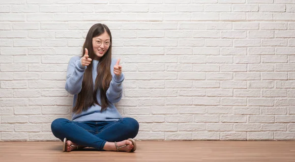 年轻的中国女人坐在砖墙上的地板上 用快乐而滑稽的脸指着镜头 良好的能量和活力 — 图库照片