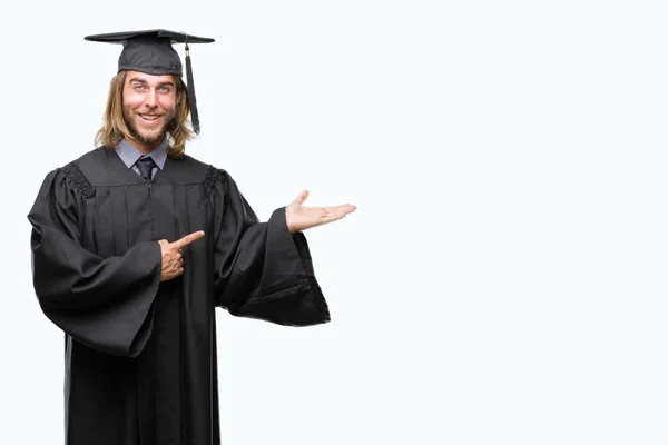 Young handsome graduated man with long hair over isolated background amazed and smiling to the camera while presenting with hand and pointing with finger.