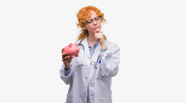 Young Redhead Doctor Woman Holding Piggy Bank Serious Face Thinking — Stock Photo, Image