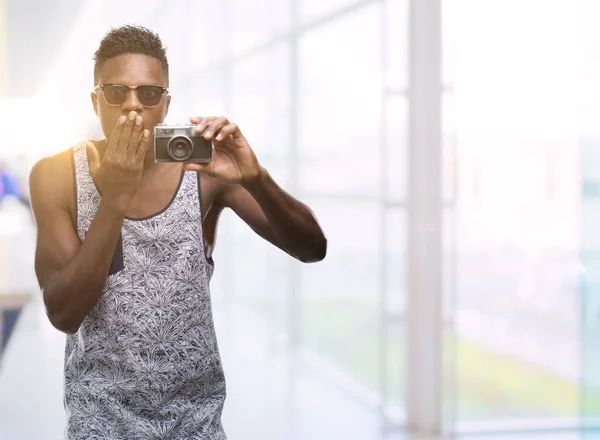 Young african american man holding vintage camera cover mouth with hand shocked with shame for mistake, expression of fear, scared in silence, secret concept