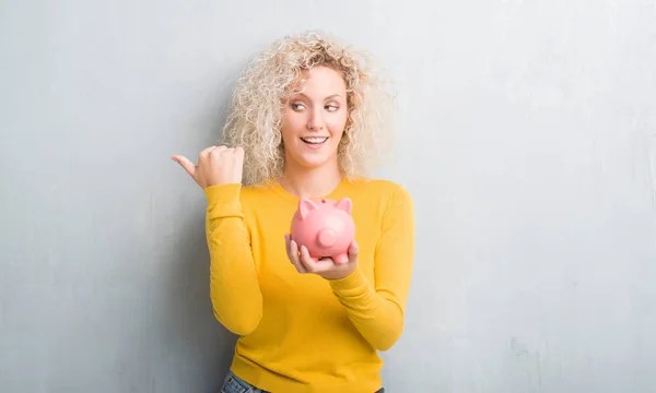 Young Blonde Woman Grunge Grey Background Holding Piggy Bank Pointing — Stock Photo, Image