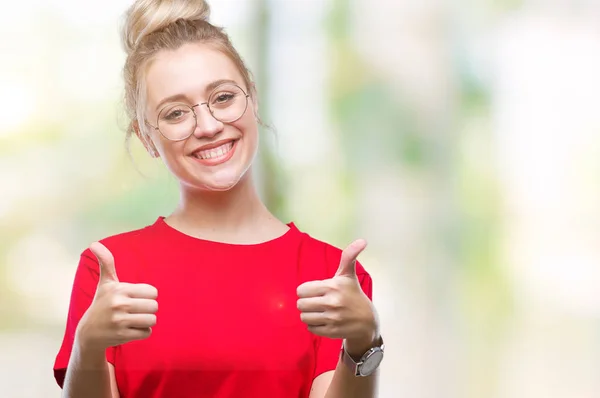 Young blonde woman wearing glasses over isolated background approving doing positive gesture with hand, thumbs up smiling and happy for success. Looking at the camera, winner gesture.