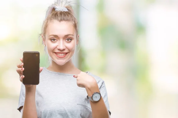 Young blonde woman showing screen of smartphone over isolated background with surprise face pointing finger to himself