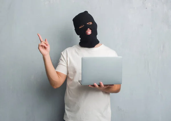 年轻的高加索黑客男子在灰色的格格墙上做网络攻击使用笔记本电脑非常高兴用手和手指指向身边 — 图库照片