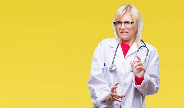 Young beautiful blonde doctor woman wearing medical uniform over isolated background disgusted expression, displeased and fearful doing disgust face because aversion reaction. With hands raised. Annoying concept.