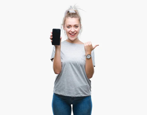 年轻的金发女子显示的智能手机屏幕上的孤立背景指向和显示与拇指到一边 快乐的脸微笑 — 图库照片